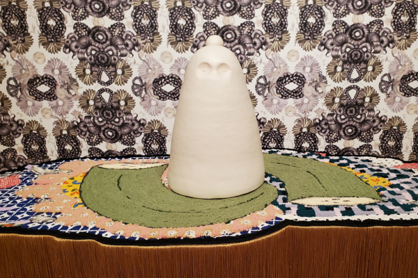 A white sculpture sits atop of a table with ornate decorations and tassels, on the wall behind is intricately-patterned black and white wallpaper. 