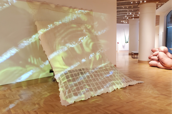 A white fabric tent is pinned to the wall and to the floor, creating a triangular shelter-like area on which videos of leaves in the sun are projected. 