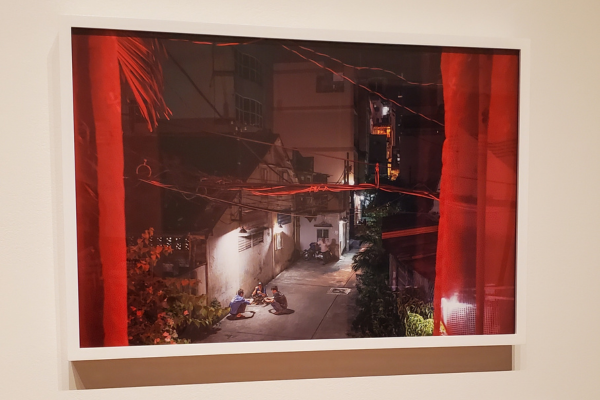 A photograph of the view out of a window, framed by red curtains, shows an alleyway at night where a trio of people play cards.